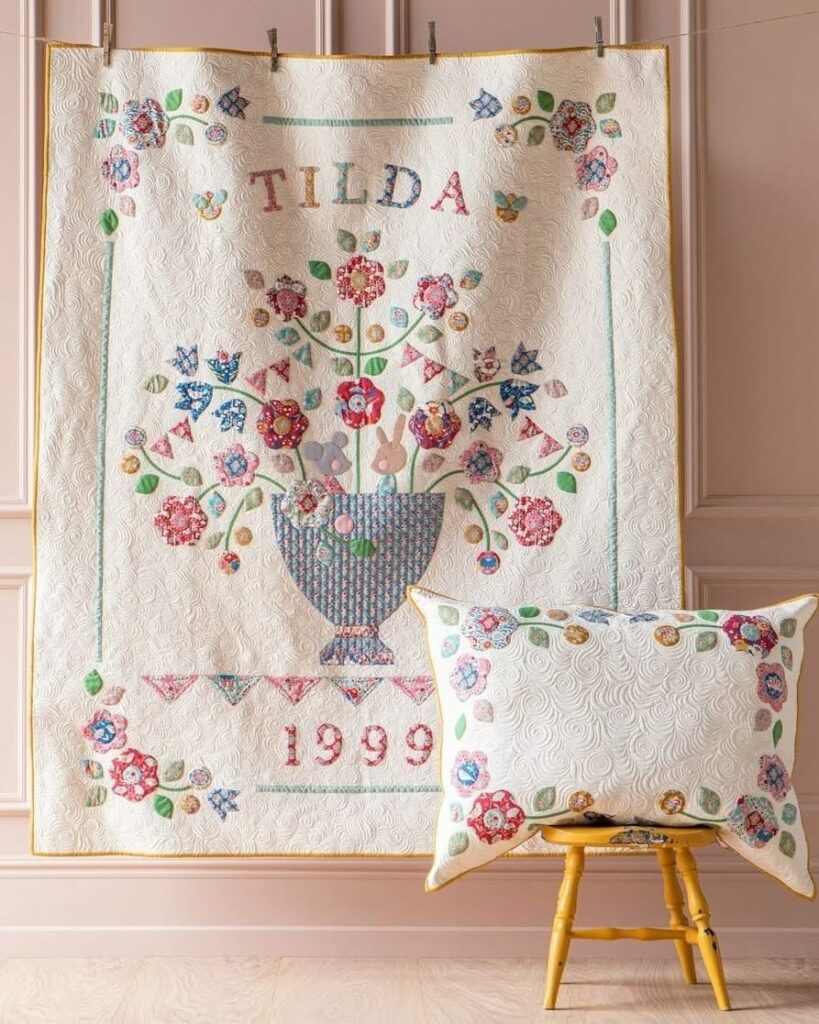 Colcha de patchwork con diseño floral en un jarrón, la palabra "TILDA" y el año "1999", acompañada de un cojín a juego sobre una silla amarilla.