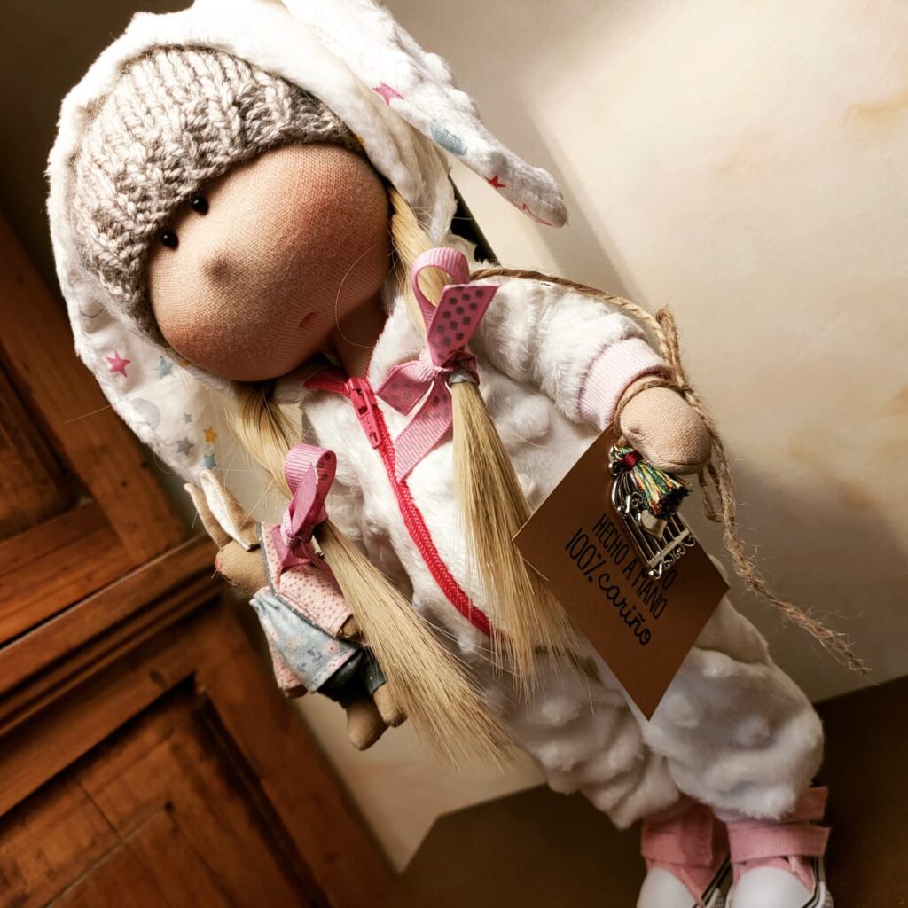 Muñeca artesanal de tela con gorro de lana, trenzas rubias y ropa blanca decorada con detalles rosas.