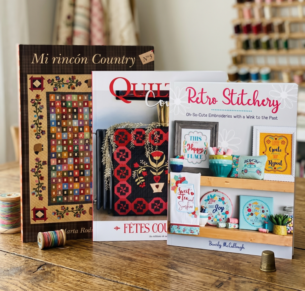 Libros de bordado y patchwork titulados "Mi rincón Country", "Quilt" y "Retro Stitchery" sobre una mesa de madera.